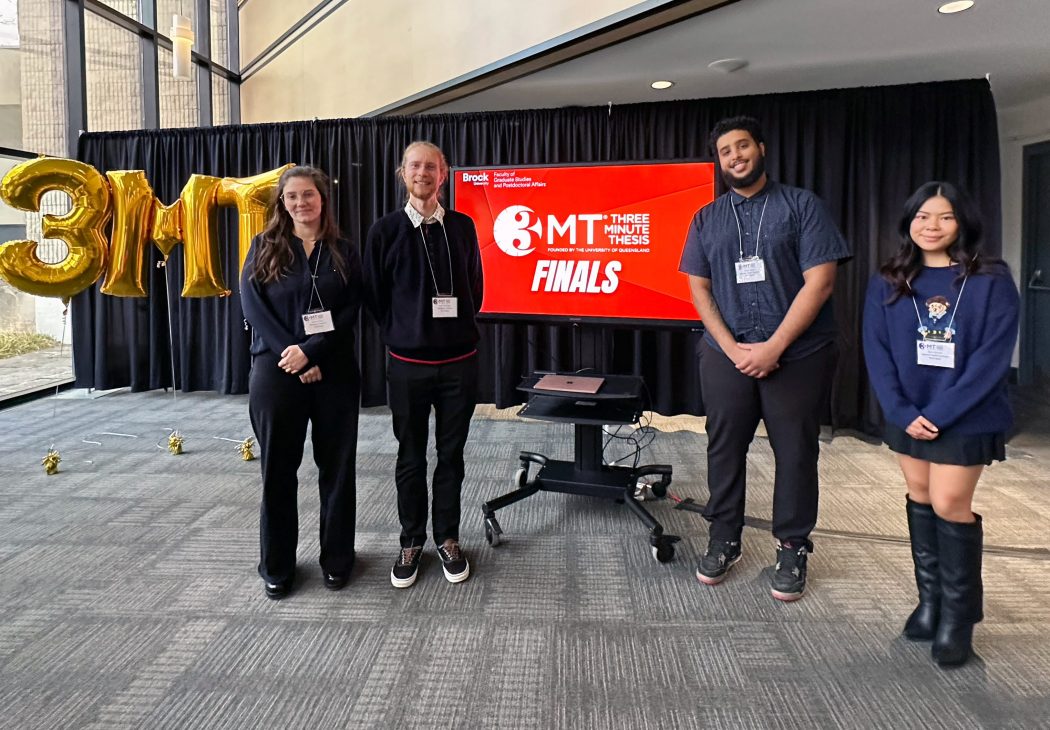 Four people stand together in front of a screen that says "Three Minute Thesis finals." 