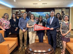A group of people pose for a photo in an event room. Three of them are holding a Brock University banner in the front.