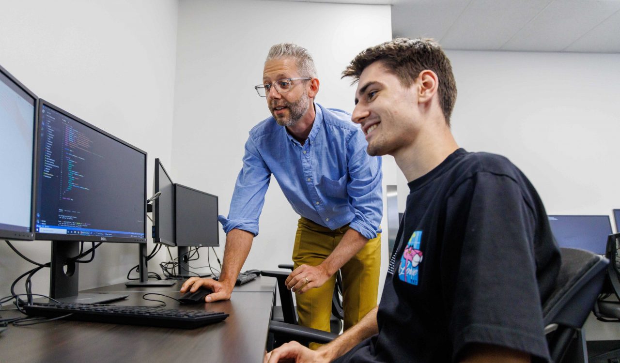 Two men look at computer monitors in an office.