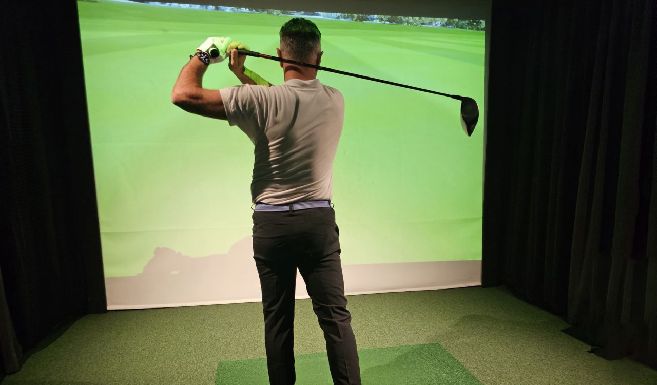 A man swings a golf club in front of a screen at at indoor golf venue.