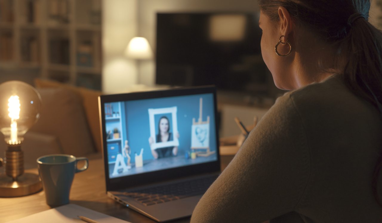 A woman sits at a desk in dark light looking at a computer screen which shows a bright blue image of a person holding a photo frame around their head.