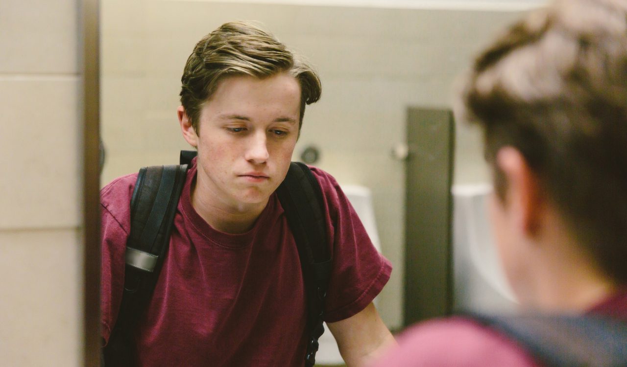 A sad teenage boy stands in front of a mirror looking at his reflection.