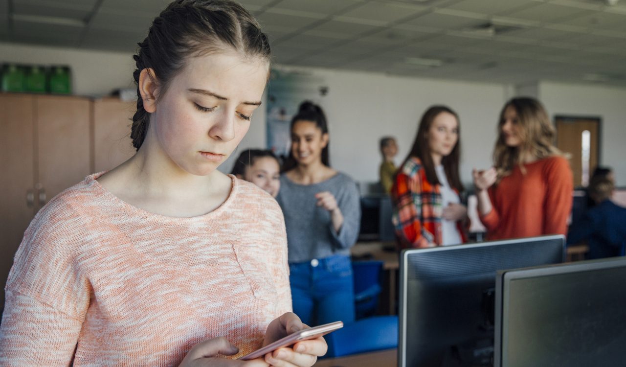 A sad teenage girl looks at her phone while classmates laugh at her in the background.