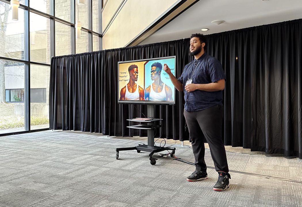 A man presents to an unseen audience with a television screen on in the background.