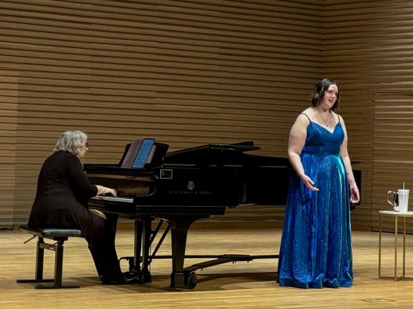 A woman in a blue dress stands on stage singing beside a woman wearing black playing the piano.