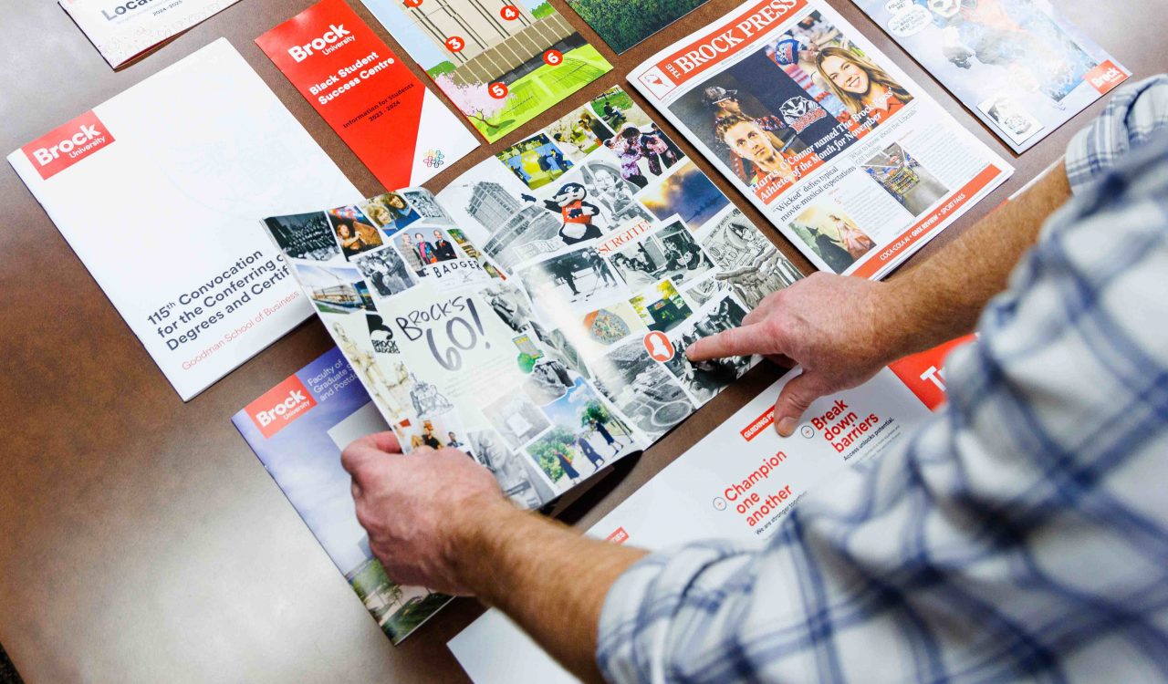 A person flips through a publication celebrating Brock’s 60-year history, surrounded by various magazines, brochures and student success resources.