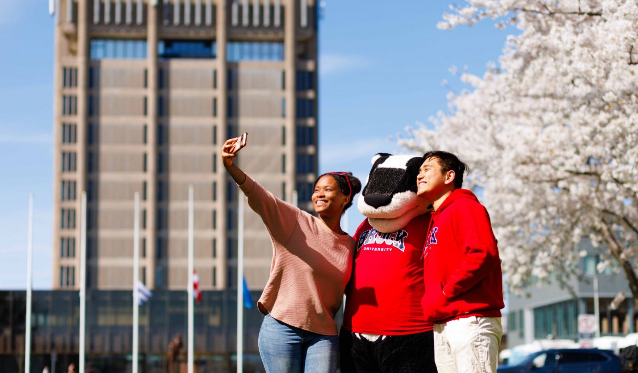 Two students and Brock University's mascot, Boomer the Badger, take a selfie under blooming cherry blossoms in front of Schmon Tower on a sunny spring day.