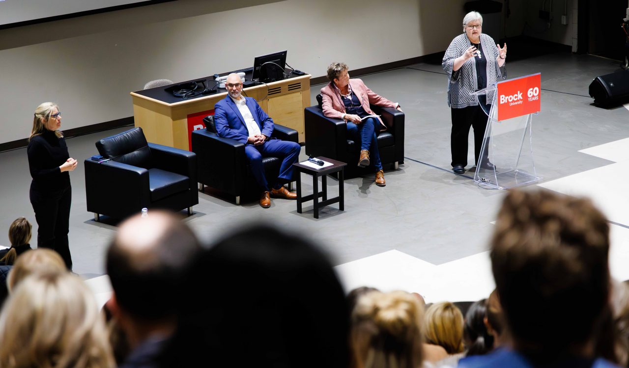 A speaker addresses the audience during a Brock University panel event focused on sustainability, as panelists and an ASL interpreter engage with attendees in a packed lecture hall.