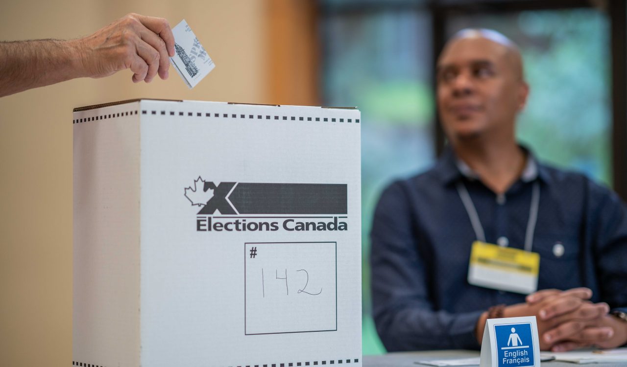 An “Elections Canada” ballot box sits in the foreground on a table with a man sitting in the background.