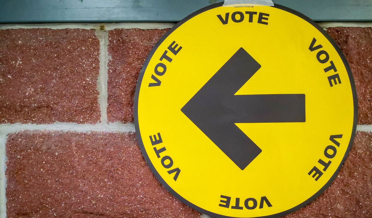 A round yellow sign with a black arrow and the word "vote" on it several times hanging on a brick wall.