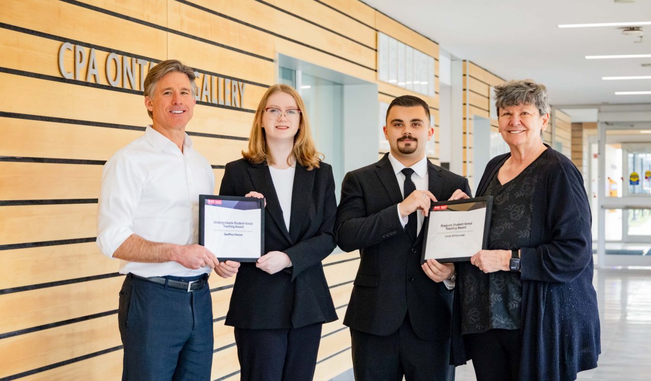 Four people pose holding two certificates between them in a hallway.