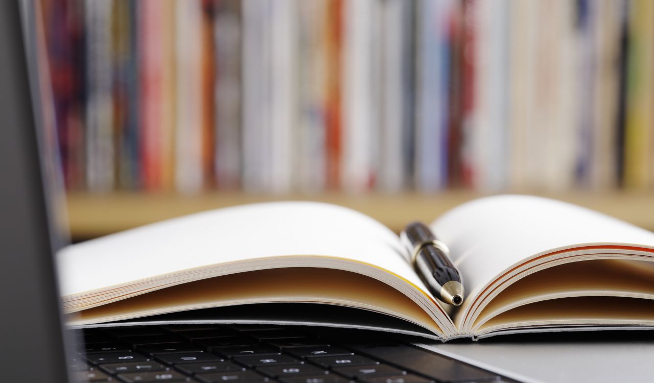 Close up of open note pad with a pen and laptop computer on a table in a library.