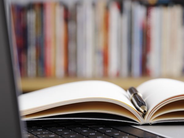 Close up of open note pad with a pen and laptop computer on a table in a library.