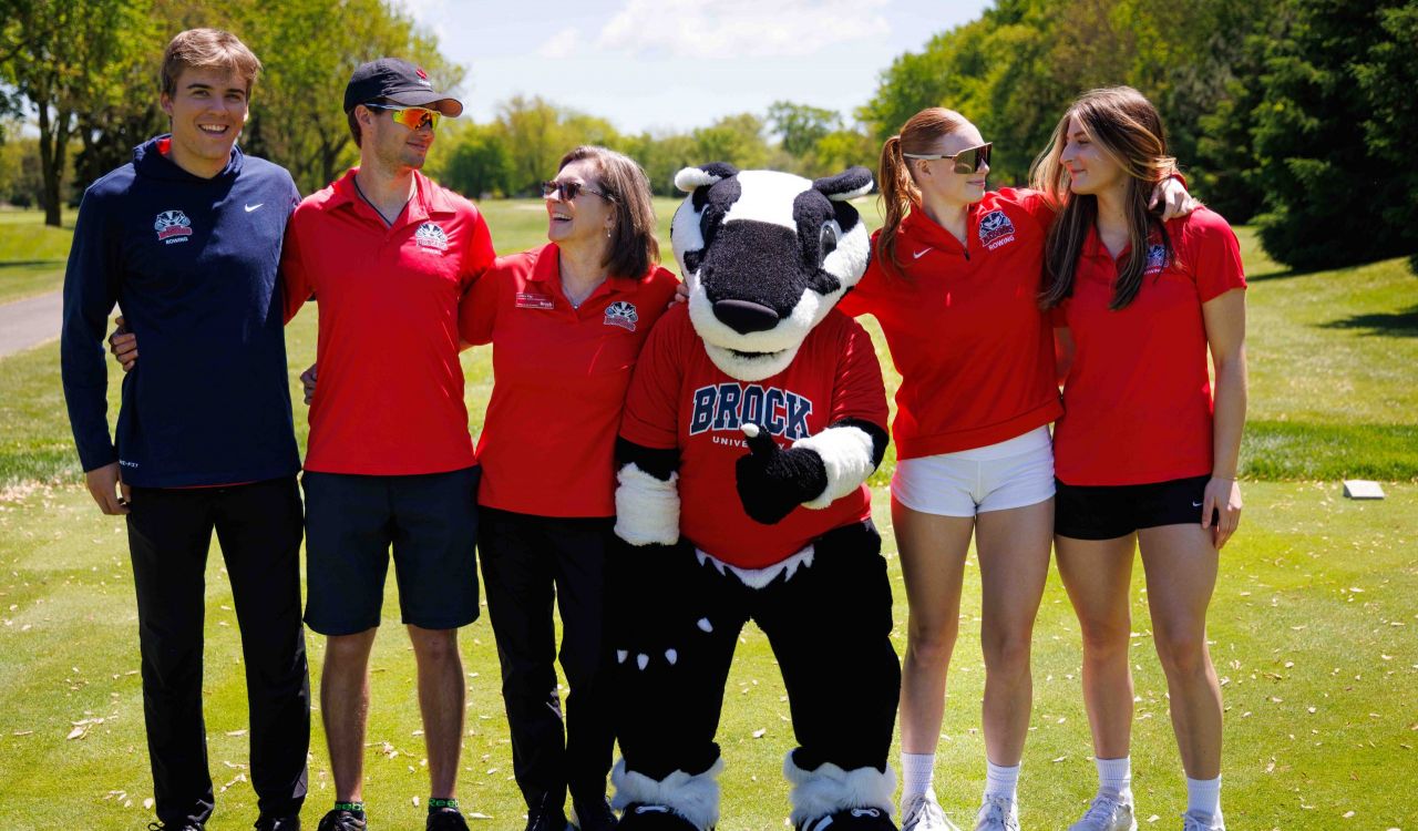 A group of people smiles while posing with a mascot on a sunny golf course.