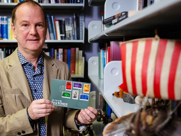 Brock Professor Andrew McDonald stands in an office lined with colourful books holding a set of colourful stamps.