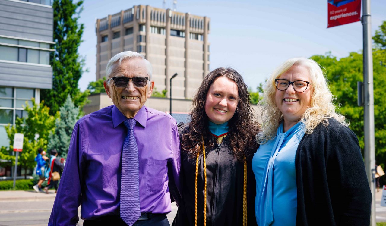 Three people stand outside, one in a Convocation gown, in front of a large concrete tower on a university campus.