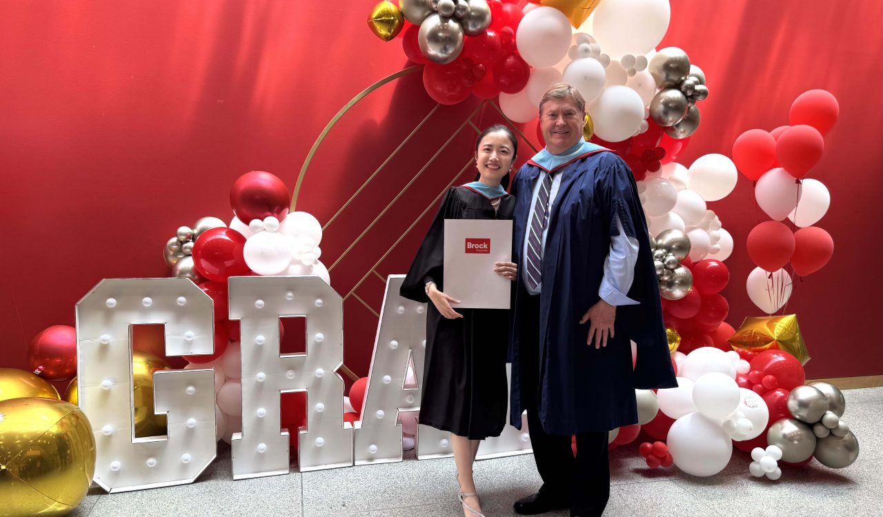 A man and woman stand next to each other wearing Convocation regalia in front of a colorful balloon backdrop.