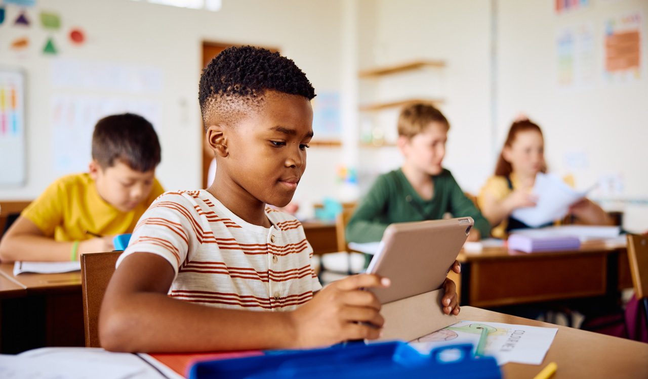 Boy sits in classroom of students and holds a tablet.