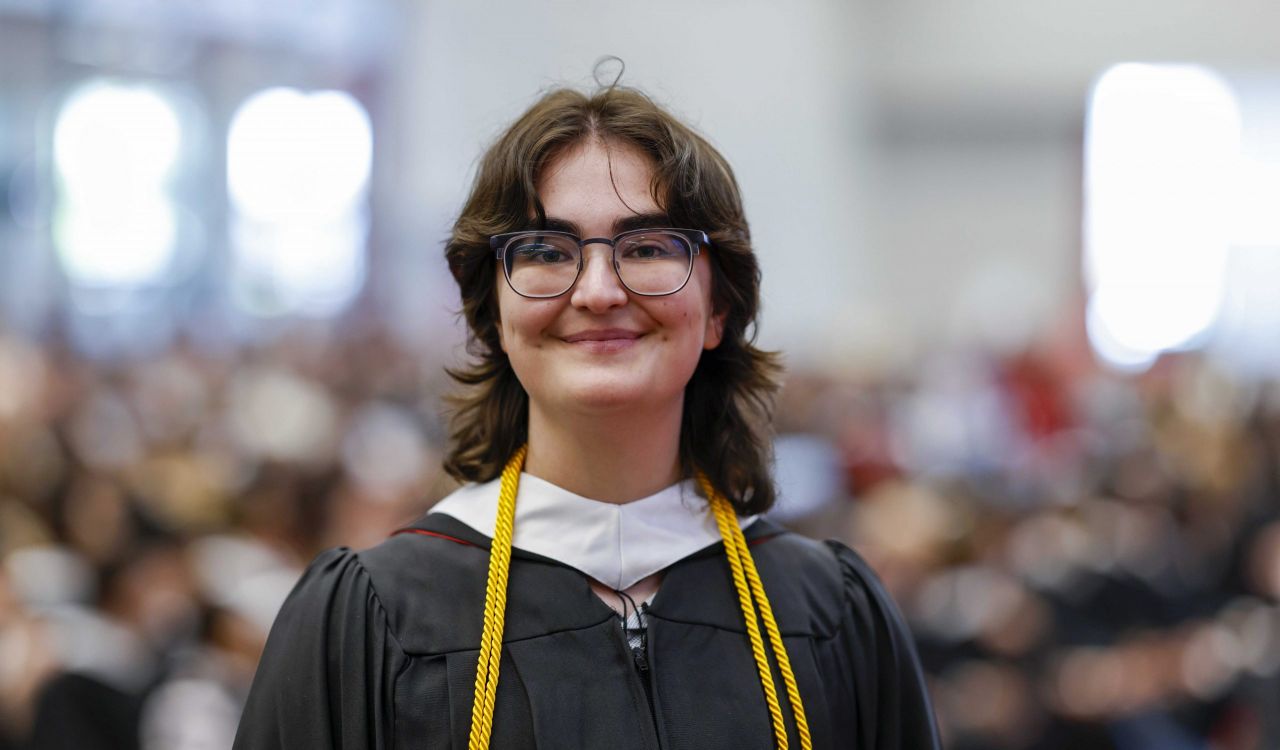 A person in an academic gown smiles while standing in a university graduation ceremony with an audience visible in the background.