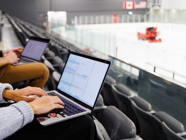 Two people work on laptops while seated in an ice hockey arena.