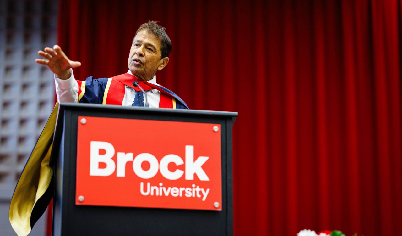 A man wearing academic robes speaks from a podium.