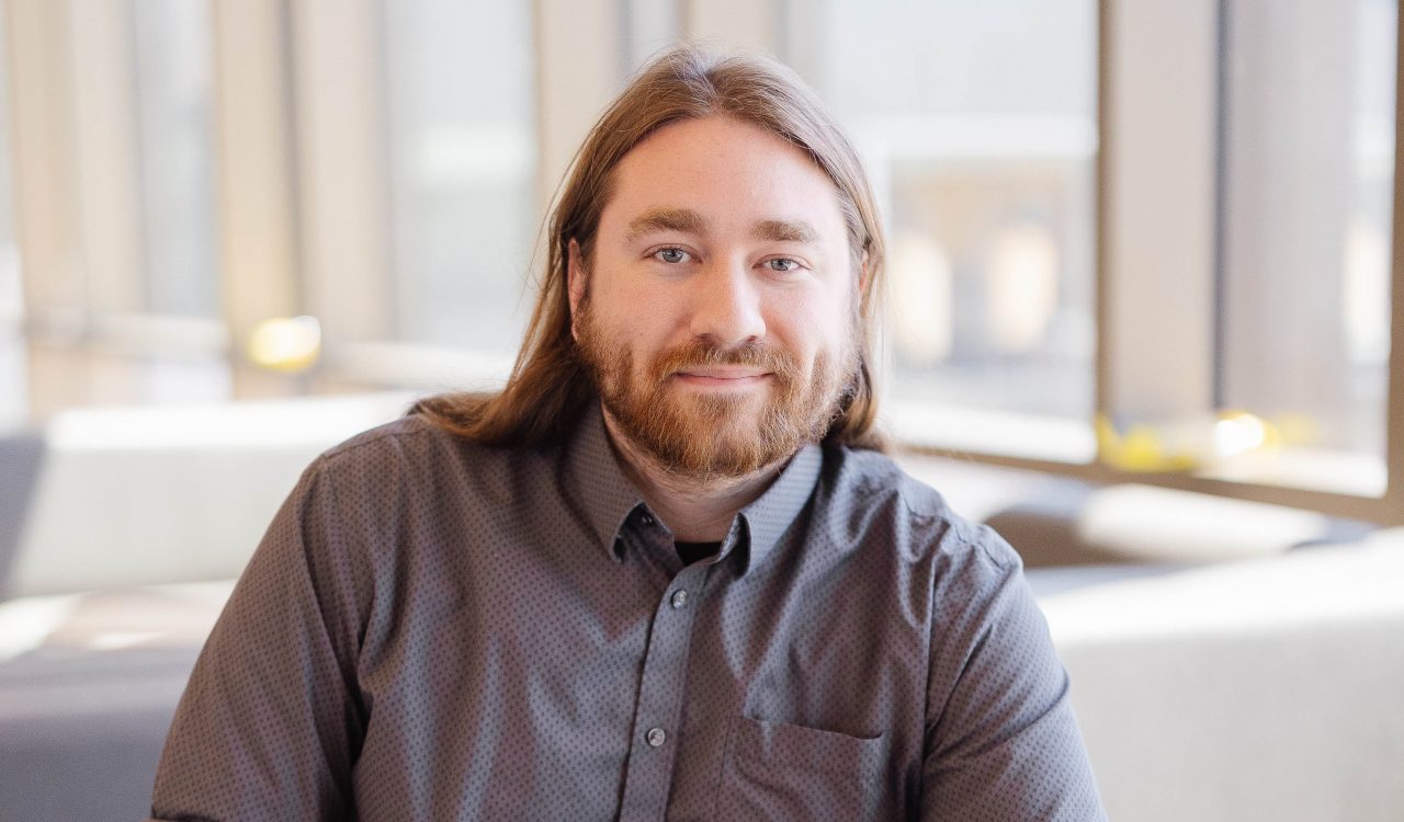 A man with long hair sits in casual business attire in a brightly lit room smiling at the camera.