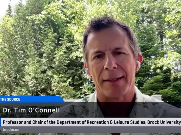 Man sits in front of trees and does a television interview.