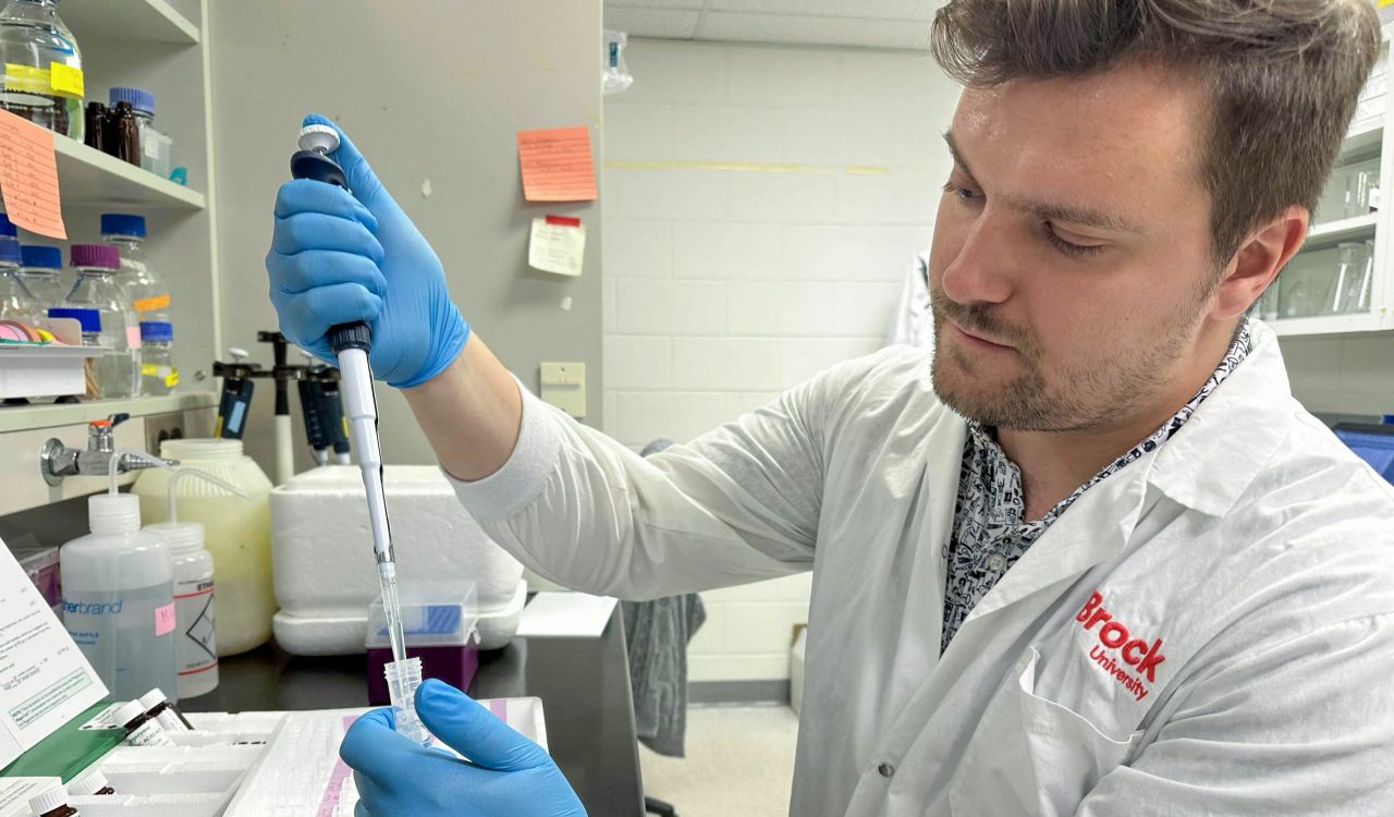 A male sits at a desk in a scientific lab, preparing samples with a liquid dispenser.