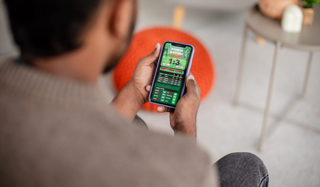 A man sits in a lounge space while looking at a sports betting app on his phone.