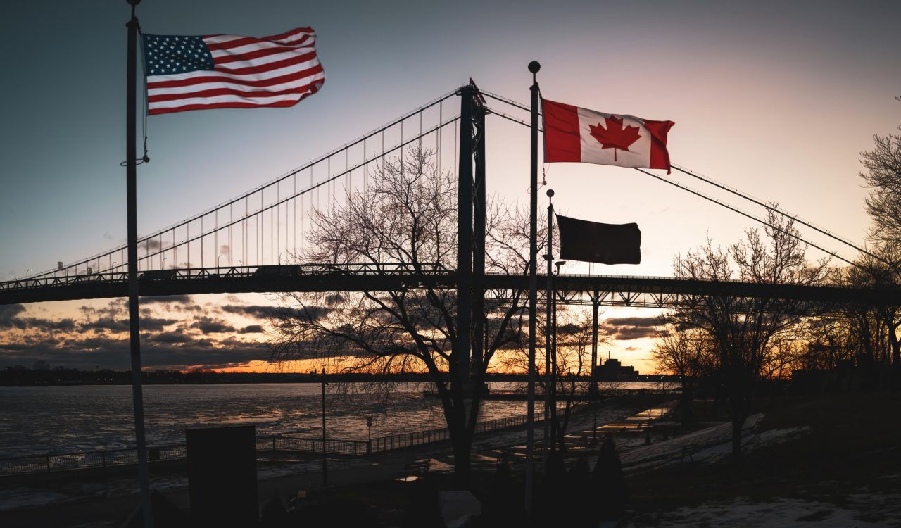 An American flag flies beside a Canadian flag with a bridge in the background.