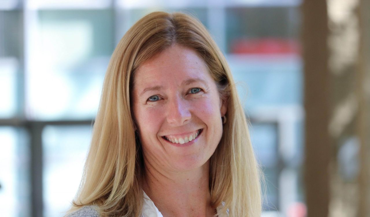 A smiling woman stands in a sunny hallway at a university.