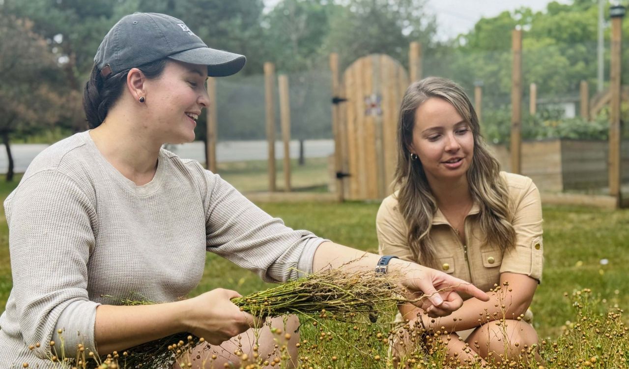 Two women sit in a field looking at flax plants.