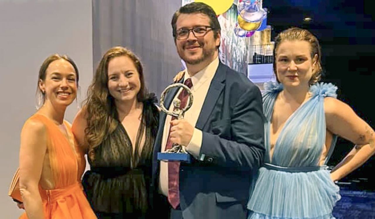 Three women and a man holding an award all dressed in formal attire stand backstage at an event.
