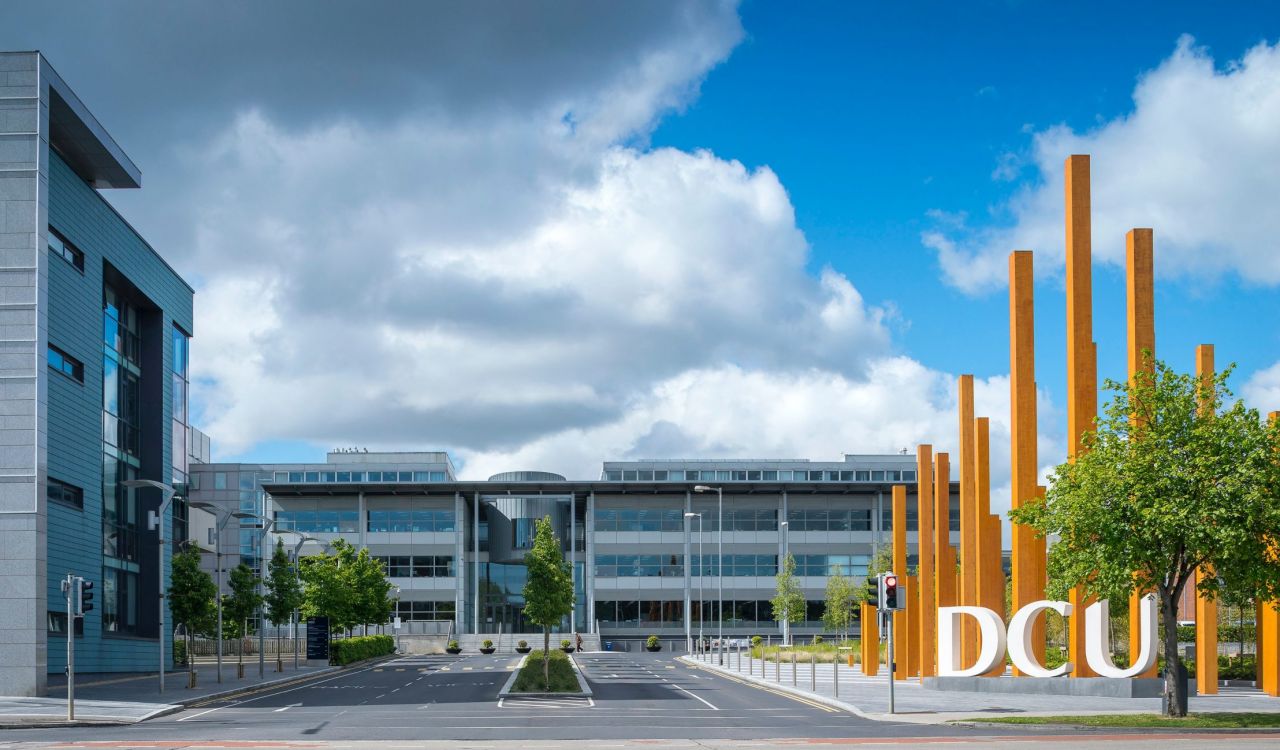 Exterior view of Dublin City University campus featuring modern glass buildings, tall orange steel sculptures, and the large DCU sign under a partly cloudy sky.