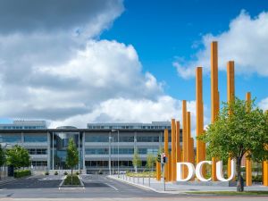 Exterior view of Dublin City University campus featuring modern glass buildings, tall orange steel sculptures, and the large DCU sign under a partly cloudy sky.