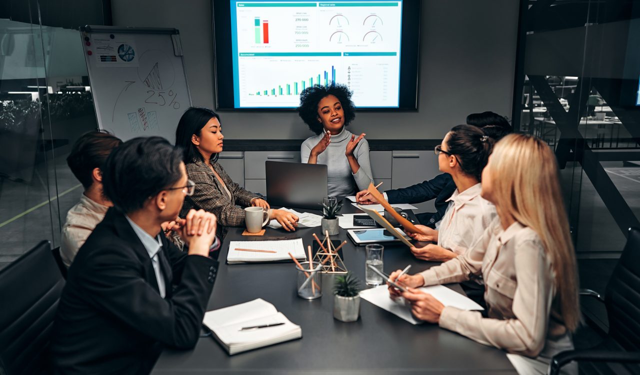 A group of people sit around a table looking at a woman speaking and gesturing, with a screen displaying graphs behind her head.