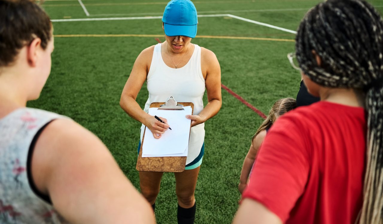 Female coach explaining game plan on a clipboard to women soccer team during practice session outside on a sports field.