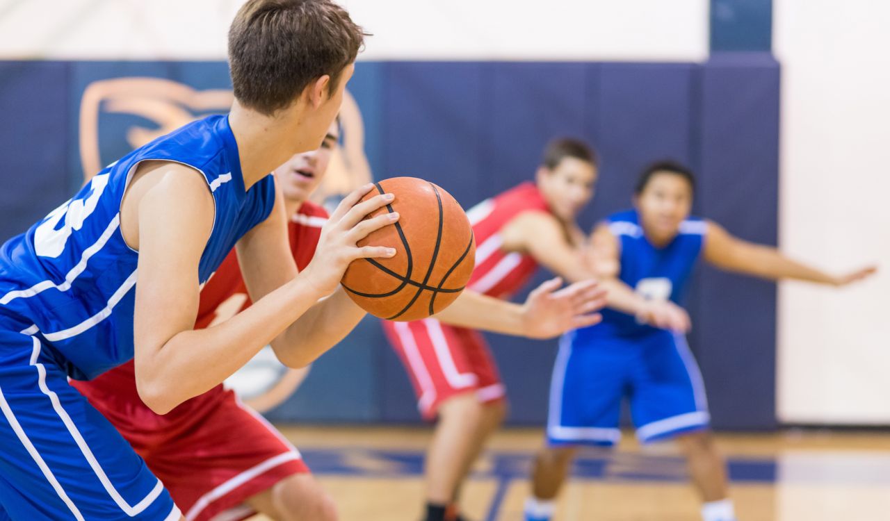Teens in jerseys play basketball on an indoor court.