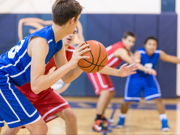 Teens in jerseys play basketball on an indoor court.