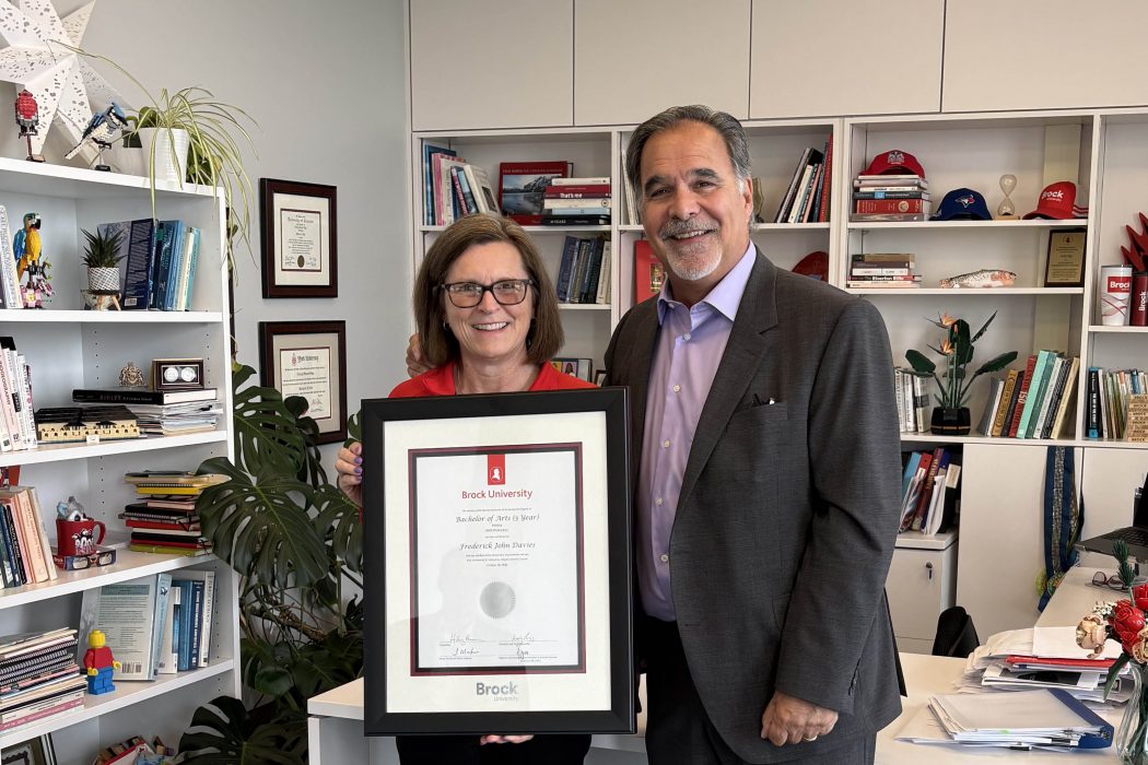 A man and a woman pose with a framed university diploma in an office.