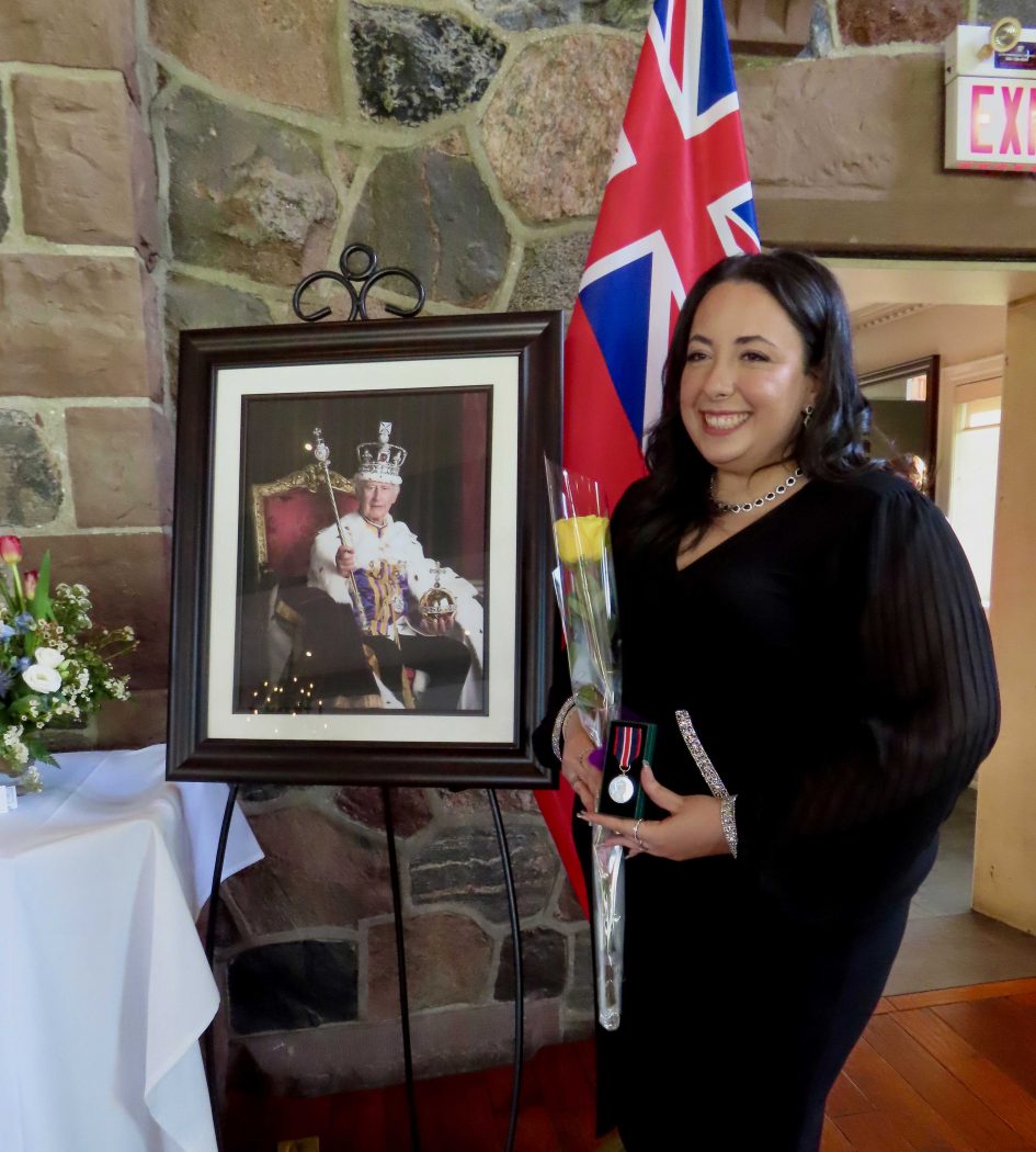 A woman wearing black holds an award certificate smiling while standing beside a portrait of King Charles III.