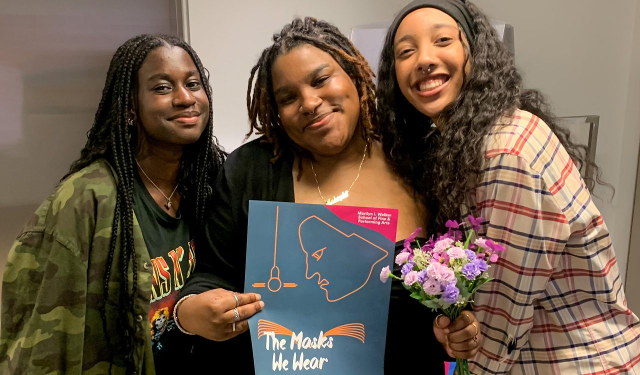 Three smiling people stand together indoors, with the person in the centre holding a poster for the Brock University Department of Dramatic Arts One Acts Festival 2025 titled 'The Masks We Wear.'