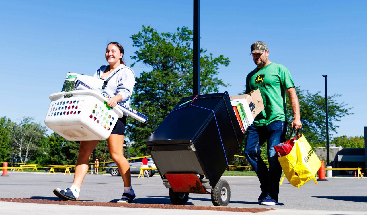A young woman smiles while carrying a white laundry basket filled with belongings, walking beside a man in a green shirt and cap who pushes a dolly loaded with a mini-fridge and boxes.