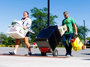 A young woman smiles while carrying a white laundry basket filled with belongings, walking beside a man in a green shirt and cap who pushes a dolly loaded with a mini-fridge and boxes.