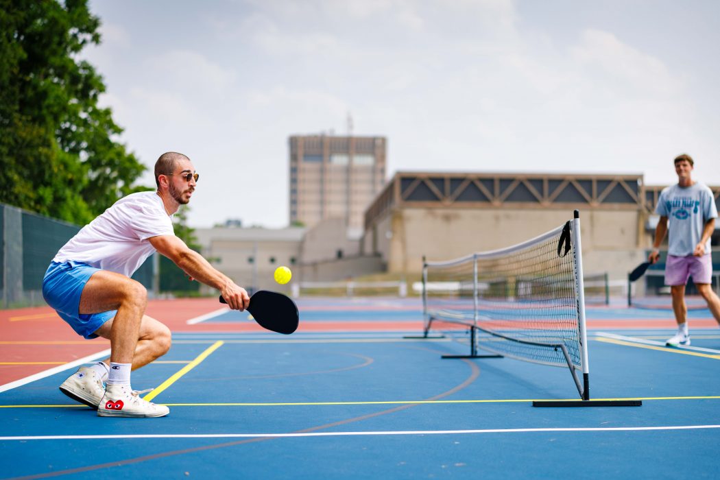 A man crouches low as he prepares to return a yellow pickleball with a paddle during a match on a bright blue outdoor court.