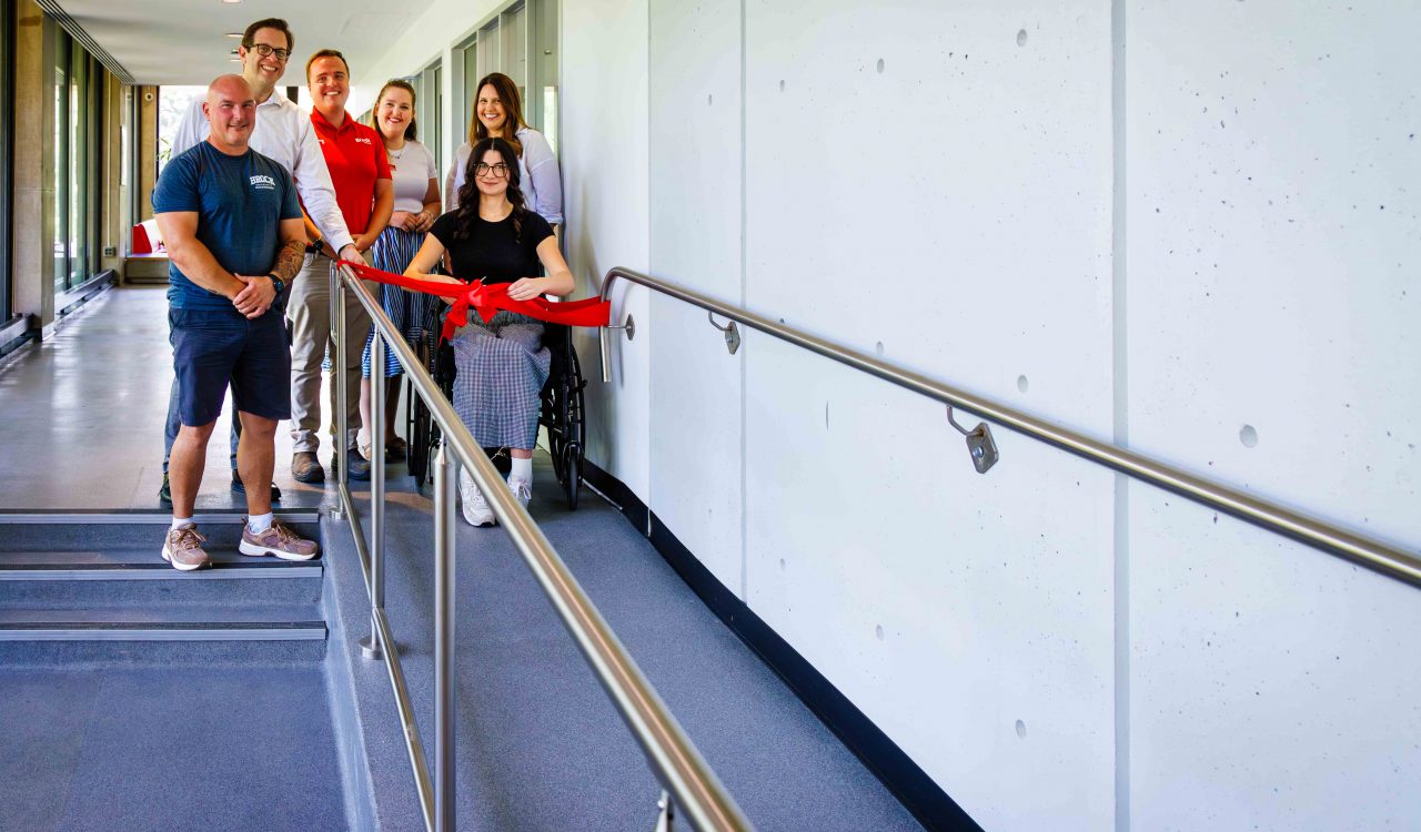 A group of six people celebrate the opening of a new wheelchair-accessible ramp. A woman seated in a wheelchair cuts a red ribbon stretched across the ramp, while others stand nearby smiling in support.