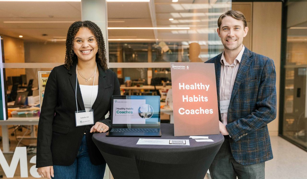 A woman and a man stand together beside a table with a poster and laptop.