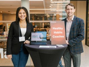 A woman and a man stand together beside a table with a poster and laptop.