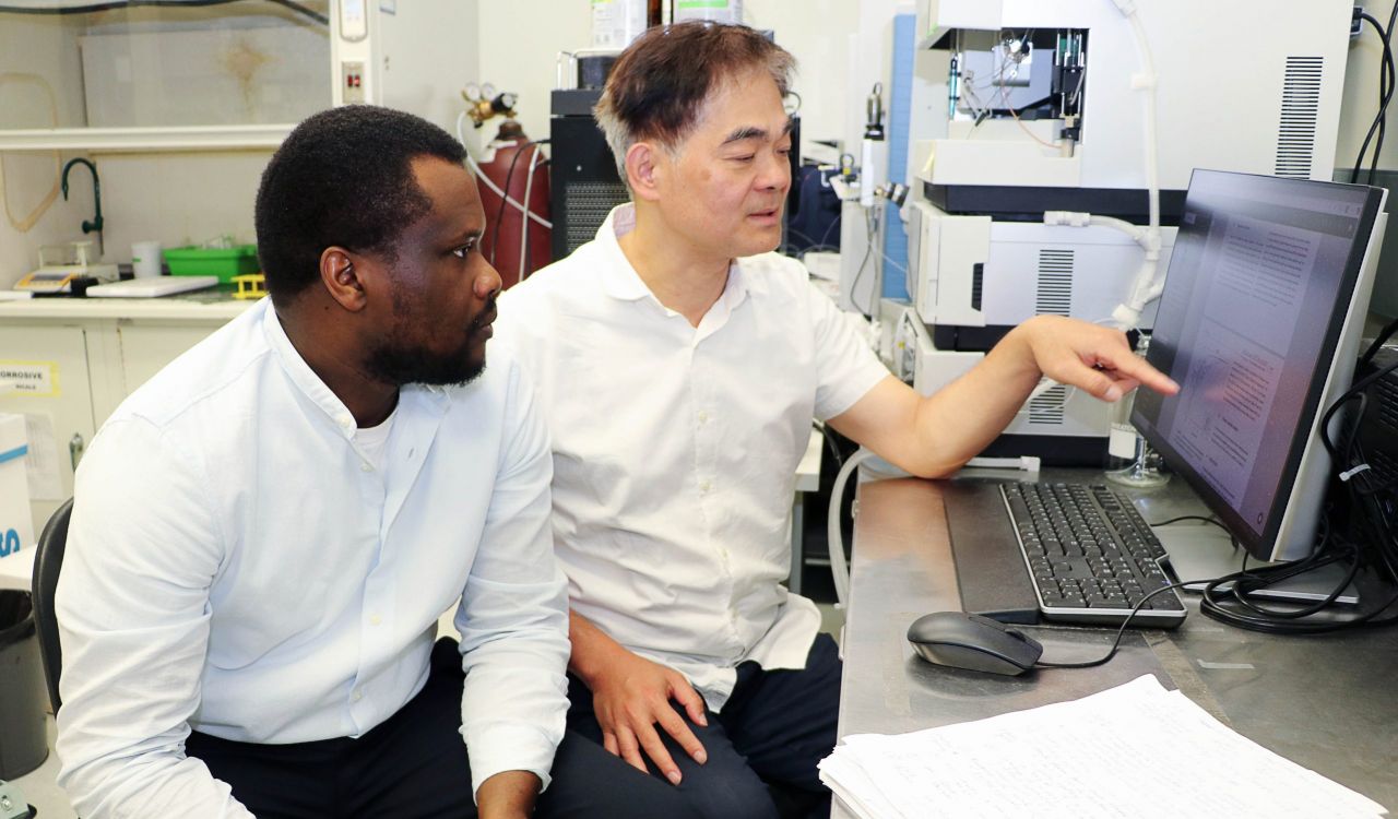 Two men sit together at a desk in a science lab looking at a computer screen.