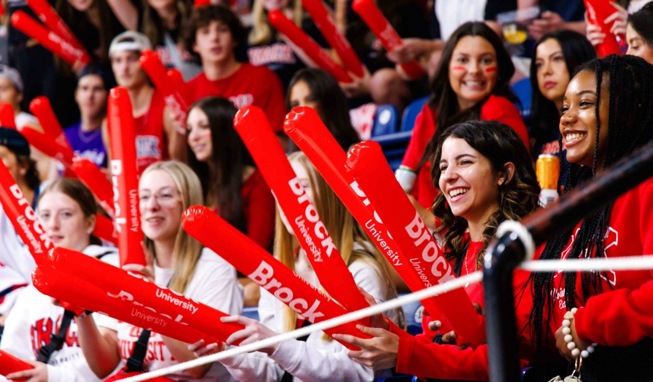 A group of enthusiastic Brock University students cheer in the stands during a sports event. They are holding bright red inflatable thundersticks branded with ‘Brock University’ while smiling and wearing red and white school spirit gear.